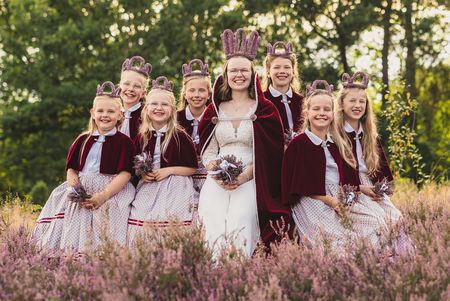 Die Heidekönigin Amelinghausen, Emma Kremer, steht gemeinsam mit ihrem sogenannten Hofstaat in der Lüneburger Heide. Alle tragen weiße Kleider und darüber einen heidefarbenen Umhang. Emma trägt zudem eine Heidekrone auf dem Kopf und einen Blumenstrauß in der Hand.