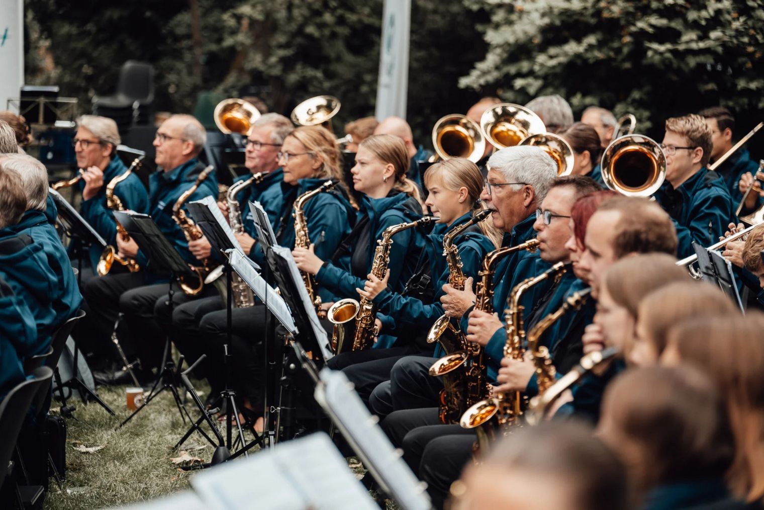 Zu sehen ist ein Blasorchester mit verschiedensten Instrumenten, wie z.B. Saxophone, Tuben, Posaunen und Trompeten. Sie tragen petrolfarbene Jacken und sitzen auf Stühlen im Gras. Vor ihnen stehen jeweils schwarze Notenständer.