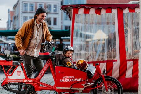 Ein Mann steht neben einem Lastenrad von "StadtRAD", in dem vorne sein Sohn sitzt. Sie stehen auf dem Marktplatz vor einem Marktstand.