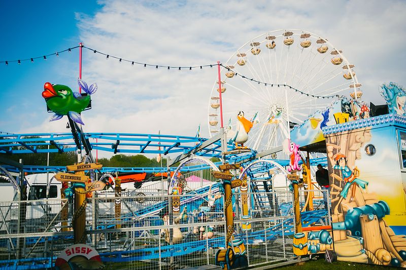 Das Bild zeigt den Frühjahrsmarkt Lüneburgs mit verschiedenen Karussell- und Fahrgeschäft-Elementen. Im Vordergrund sieht man eine bunt gestaltete Kinderachterbahn mit Comic-Figuren, Wegweisern und Dekorationen. Dahinter ragt ein großes Riesenrad in den Himmel, dessen Gondeln im Sonnenlicht leuchten. Lichterketten spannen sich über das Gelände, während der klare Himmel eine fröhliche, lebendige Festatmosphäre unterstreicht.