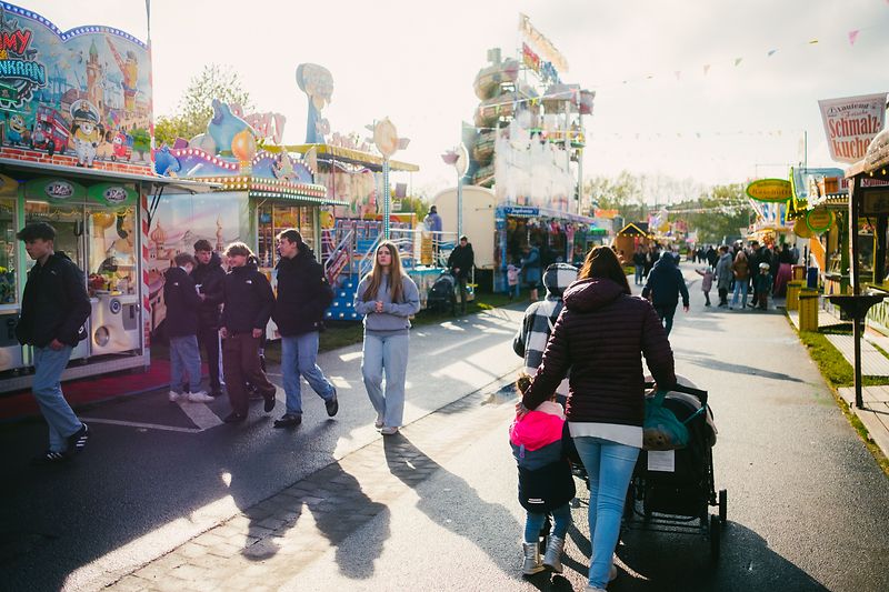 Im Vordergrund sind Personen zu sehen, wie Familien, die zwischen den Ständen des Lüneburger Frühjahrsmarkts gehen. Drumherum sieht man verschiedene Stände und im Hintergrund auch einige Fahrgeschäfte.