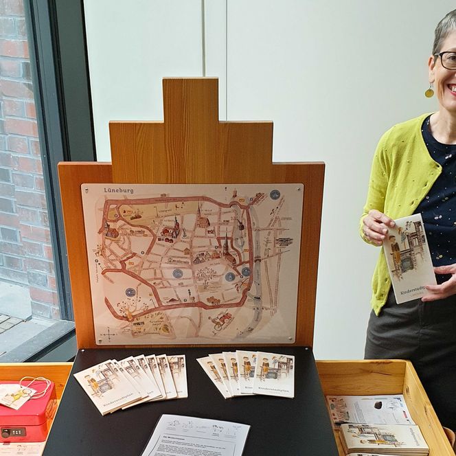 Eine Frau steht neben einem sogenannten Giebeltisch im Eingangsbereich des Museums Lüneburg. Auf diesem Tisch liegen Stadtpläne und Rätsel.
